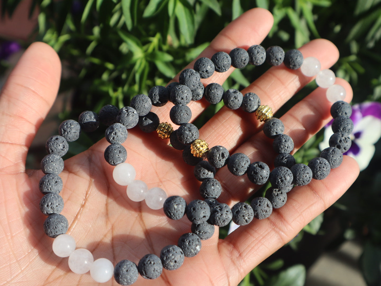Hand holding a stack of gray and white beaded bracelets with a blurred green plant background