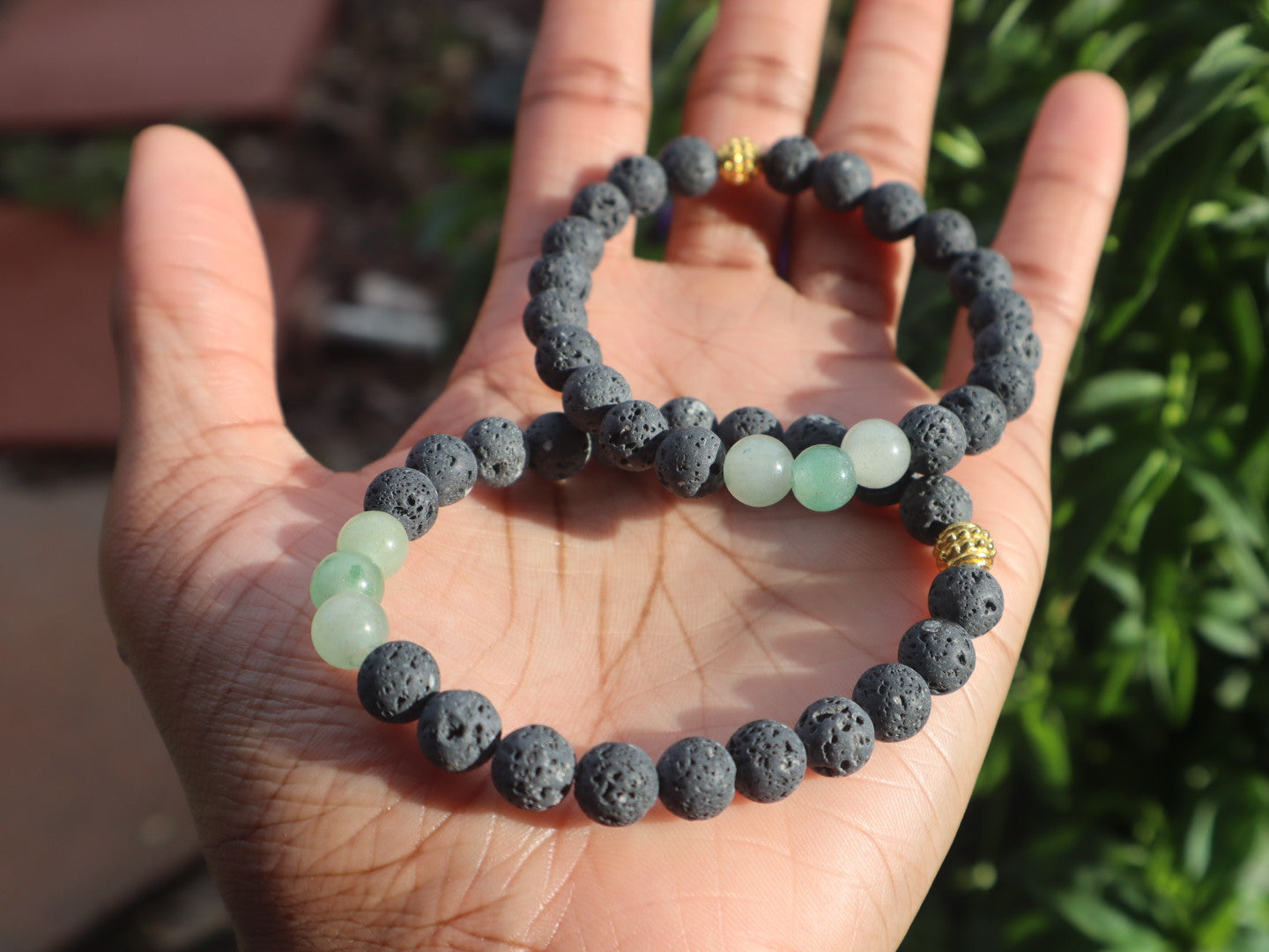 Two beaded bracelets, one with dark gray beads and the other with green beads, held in a hand against a natural background.