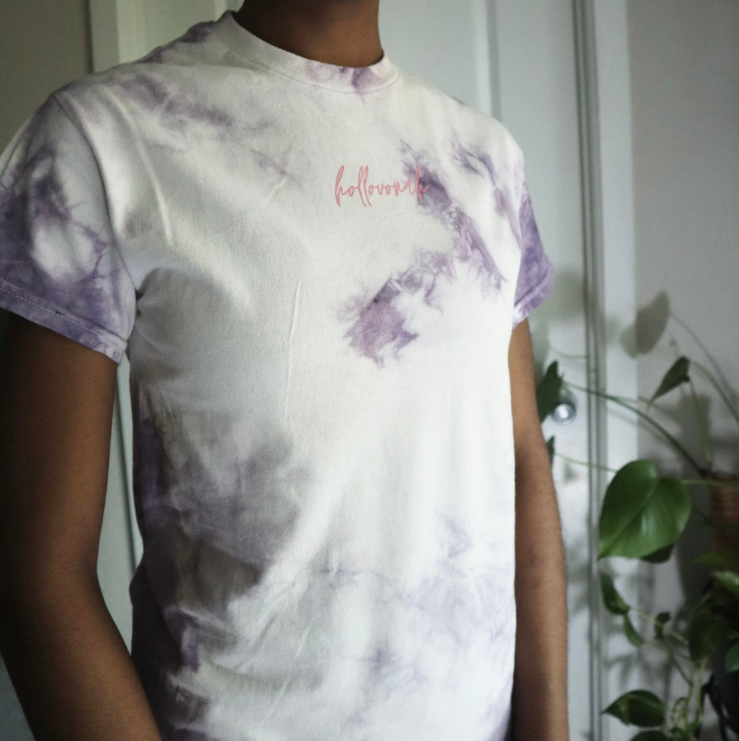 Person wearing a tie-dye t-shirt with 'hollovonah' printed on it, standing indoors with plants in the background.