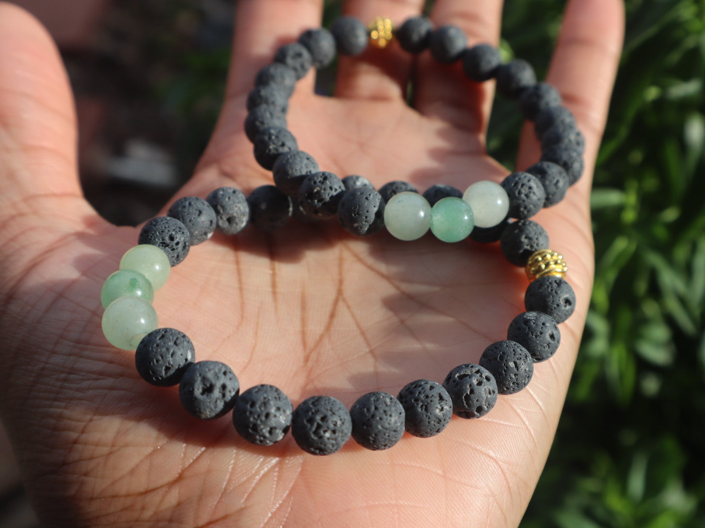 Two beaded bracelets, one with dark beads and the other with green beads, held in a hand against a blurred natural background.