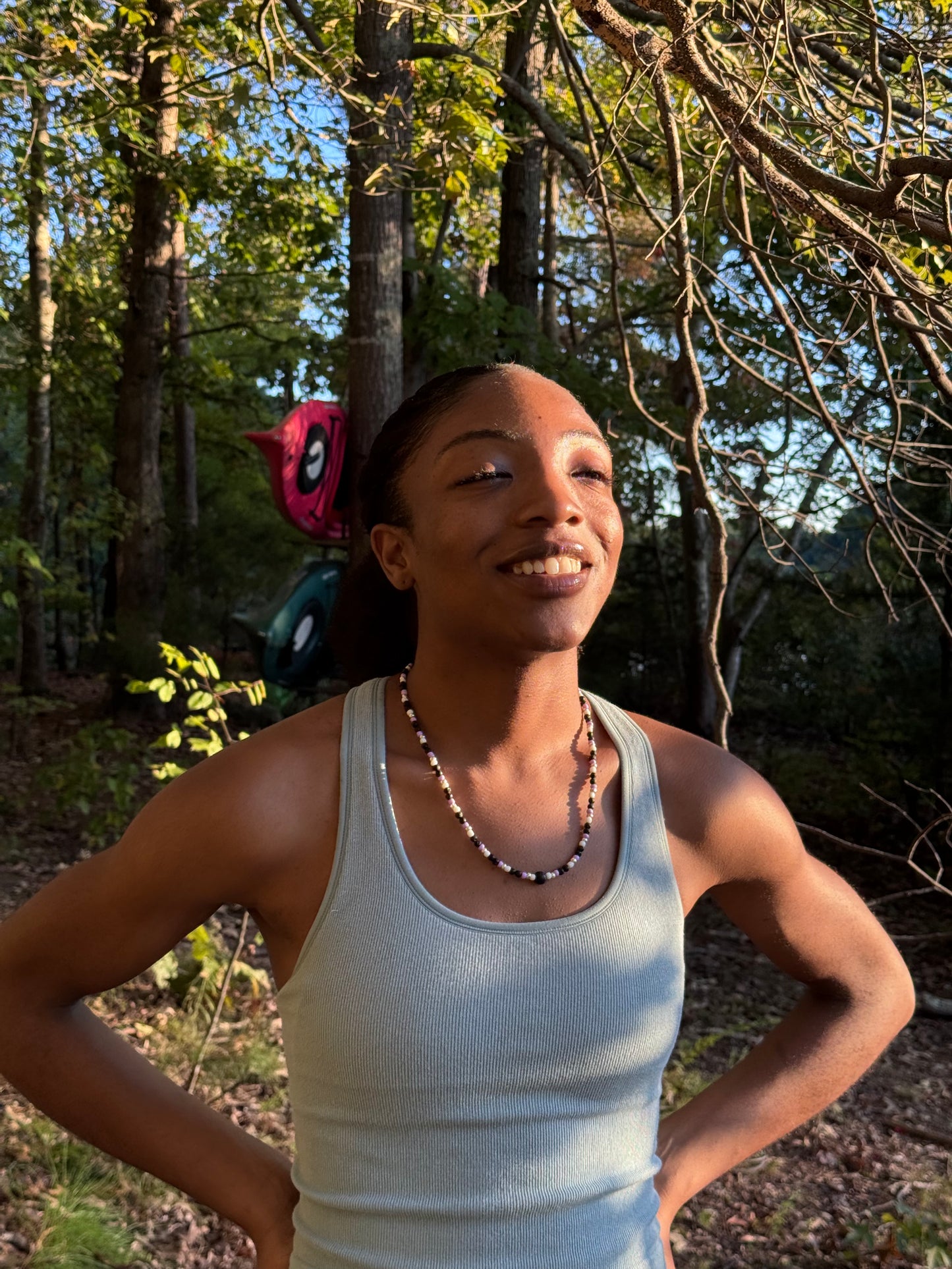 Person in a white tank top standing outdoors with trees in the background