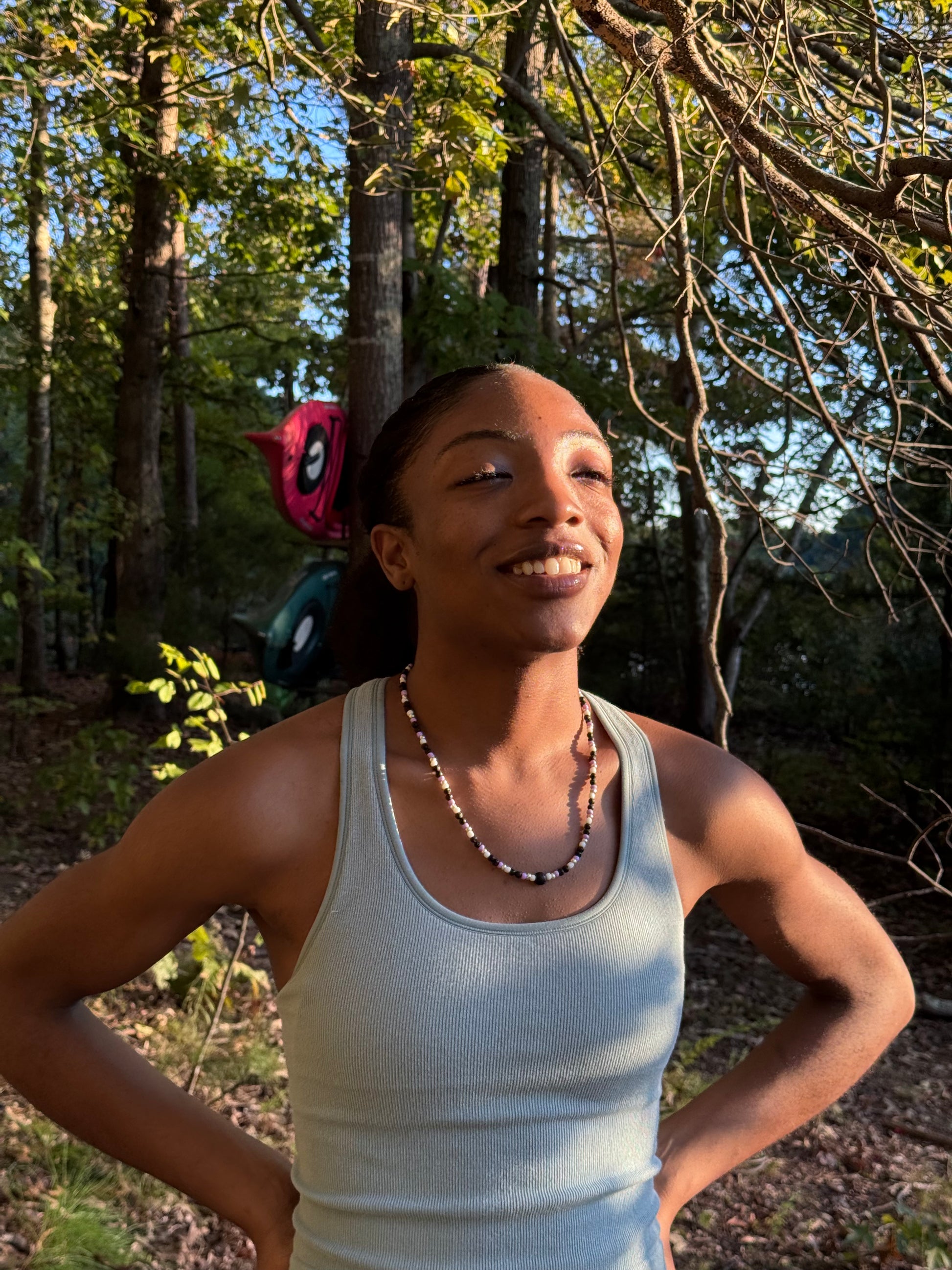 Person in a white tank top standing outdoors with trees in the background