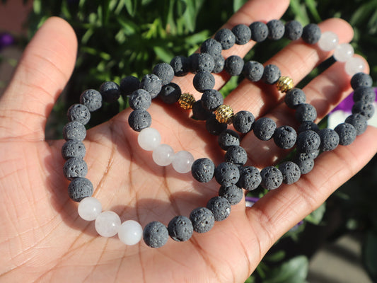 Hand holding two beaded bracelets, one black and gray, one with gold accents, against a blurred green background.