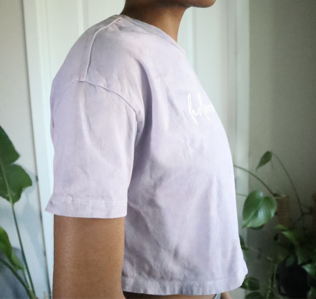 Person wearing a light purple t-shirt with plants in the background