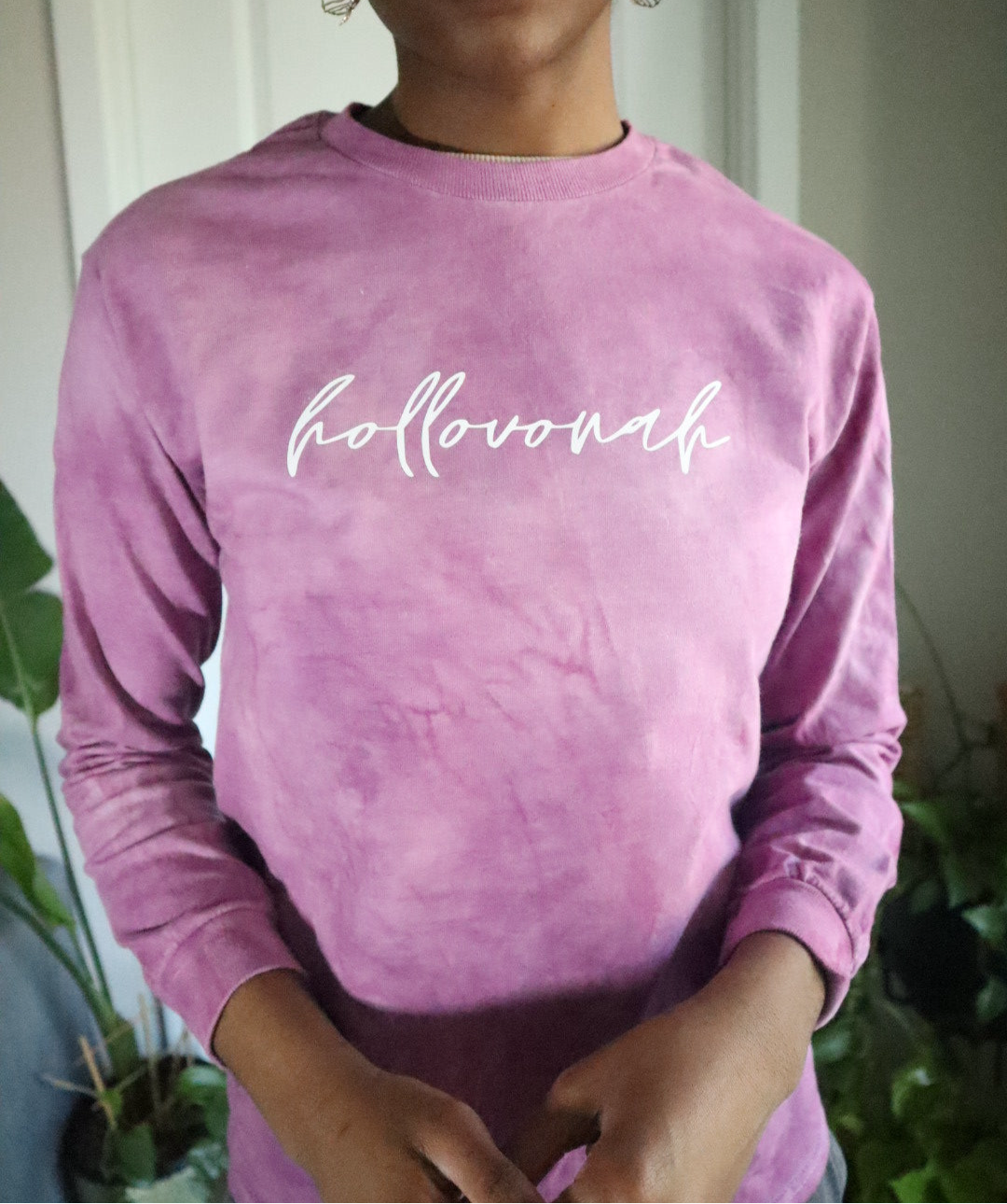 Person wearing a pink long-sleeve shirt with 'hollovonah' text, standing indoors with plants in the background.