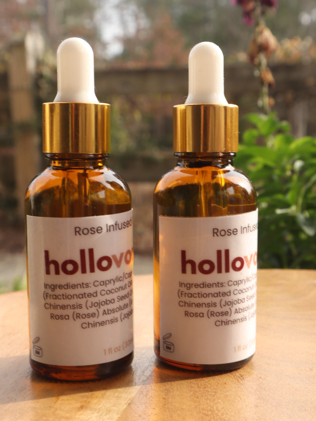 Two bottles of hollovoanh Rose Infused Oil on a wooden surface with a blurred natural background.