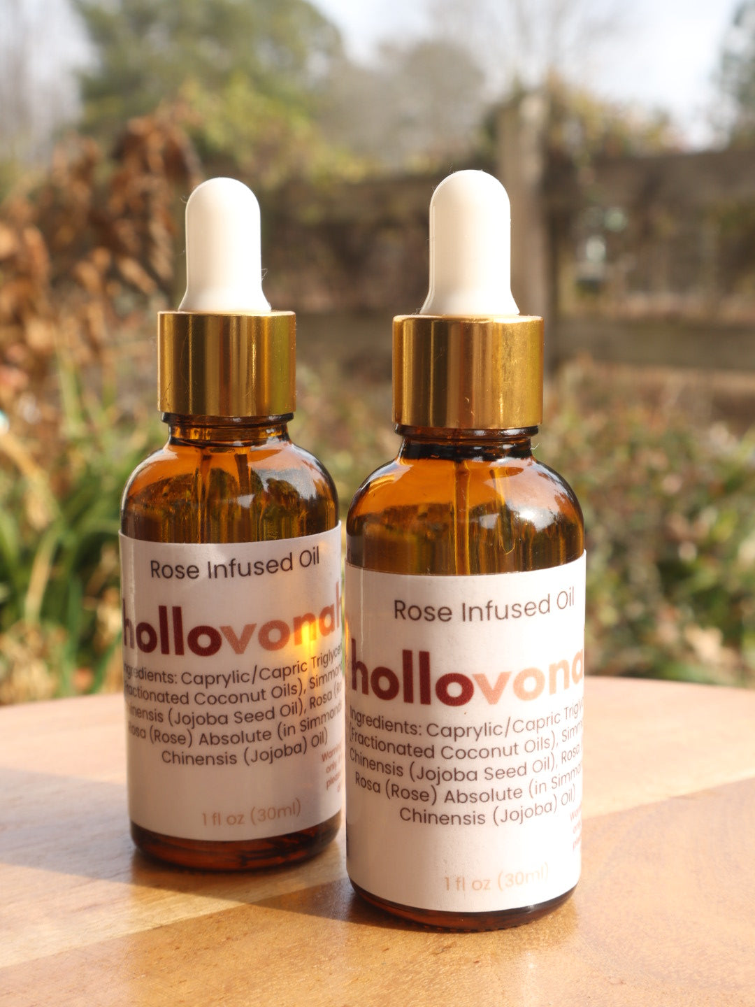 Two bottles of Hollovanh Rose Infused Oil on a wooden surface with a blurred natural background.
