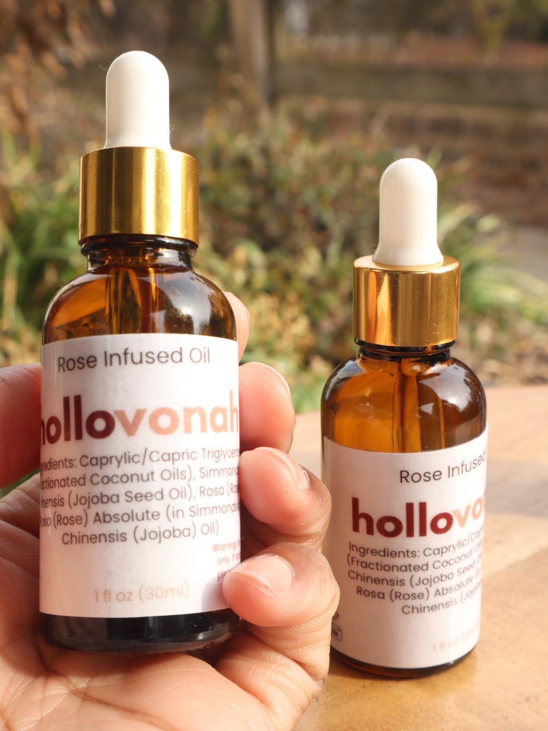 Two bottles of hollovonah Rose Infused Oil held by a hand with a blurred natural background.